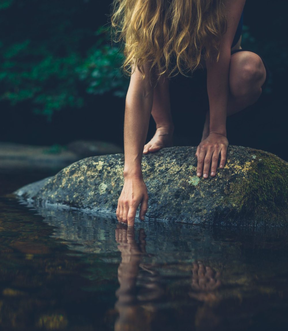 A young woman is sitting on a rock in the river and is touching the water